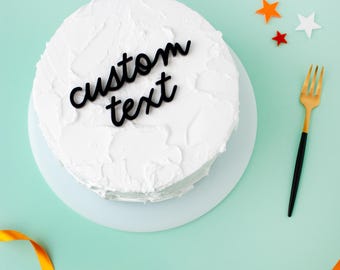 May include: A round cake covered in white frosting with a black script cake topper that reads custom text. The cake is accompanied by a gold and black fork and small orange and white star-shaped decorations on a light green surface.