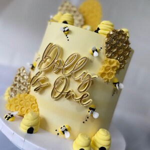 May include: A yellow cake decorated with a gold 'Dolly is One' topper, honeycomb details, and small bee figurines.