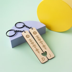 May include: Two wooden keyrings with a heart cut out of the bottom. The keyrings are engraved with a music note and a barcode. The keyrings are connected by a black chain. The keyrings are on a light green surface.
