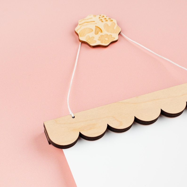 May include: A wooden sign with a scalloped edge and a white background. The sign is hanging from a white string attached to a wooden floral design.