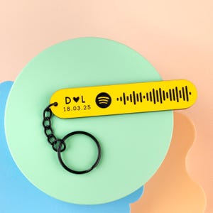 May include: A yellow keychain with a black chain and ring. The keychain has a Spotify code and the text "D ♥ L 18.03.25" in black. The keychain is on a light green and peach background.