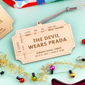 以下が含まれることがあります： 「THE DEVIL WEARS PRADA」の文字とイベント詳細が書かれた、チケット型の木製オーナメント。吊り下げ用の灰色の紐が付いており、電飾などお祝いの飾りで囲まれています。