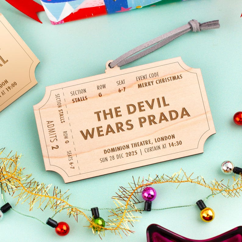 以下が含まれることがあります： 「THE DEVIL WEARS PRADA」の文字とイベント詳細が書かれた、チケット型の木製オーナメント。吊り下げ用の灰色の紐が付いており、電飾などお祝いの飾りで囲まれています。