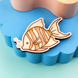 May include: Wooden fish-shaped decorative item with a striped pattern in shades of brown and tan. The fish has a dark outline and is placed on a blue, cloud-shaped surface. The item is likely a brooch, pin, or decorative element.