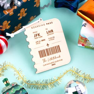 May include: A wooden boarding pass with the text "BOARDING PASS" and details including "JFK NEW YORK" and "LHR LONDON". The pass includes a flight number, date, and time. The text "The Whiteheads OUR FAVOURITE ADVENTURE 2025" is also present.