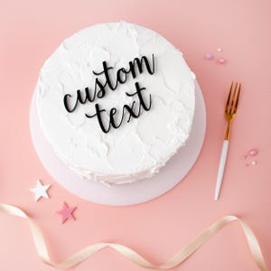 May include: A round white frosted cake with the words "custom text" in black script. A gold fork with a white handle and decorative stars and ribbon on a pink background.