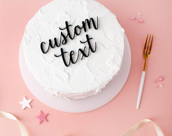 May include: A round cake covered in white textured frosting with a black script cake topper that reads custom text. The cake is placed on a white circular base next to a gold and white fork, small star-shaped decorations, and a light ribbon.