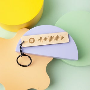May include: A wooden keychain with a Spotify code engraved on it. The keychain is attached to a black key ring and is sitting on a purple and green background.