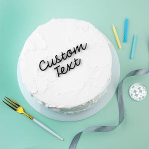 May include: A round cake covered in white frosting with a black script cake topper that reads Custom Text. The cake is accompanied by a gold-handled fork, three thin birthday candles in yellow and blue, a silver ribbon, and a disco ball icon.