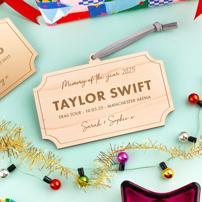 Puede incluir: Un adorno de madera con el texto "Memory of the year 2025" y "TAYLOR SWIFT". Incluye el texto "ERAS TOUR - 10.05.25 - MANCHESTER ARENA" y los nombres "Sarah + Sophie". El adorno tiene una correa de cuero gris.