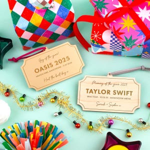 Puede incluir: Se muestran placas de madera con textos grabados como "OASIS 2025" y "TAYLOR SWIFT". Est&aacute;n adornadas con elementos decorativos como papel de regalo, guirnaldas de luces y decoraciones de fiesta. El tema son recuerdos de eventos.