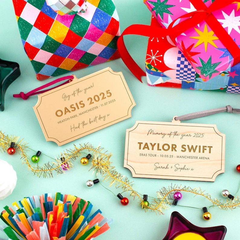Puede incluir: Se muestran placas de madera con textos grabados como "OASIS 2025" y "TAYLOR SWIFT". Est&aacute;n adornadas con elementos decorativos como papel de regalo, guirnaldas de luces y decoraciones de fiesta. El tema son recuerdos de eventos.