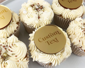 May include: Gold cupcake toppers with the text "Custom Text" on them. The toppers are placed on top of cupcakes with white frosting and gold sprinkles.