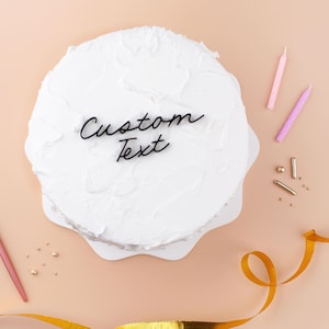 May include: A round cake covered in white frosting with a black script cake topper that reads Custom Text. The cake is surrounded by two thin pink candles, small gold metallic sprinkles, and a gold ribbon on a light peach-coloured surface.