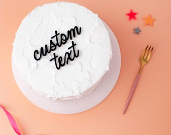 May include: A round cake covered in white frosting with a black script cake topper that reads custom text. The cake is placed on a white circular base next to a gold and pink fork, three small star-shaped decorations, and a pink ribbon.