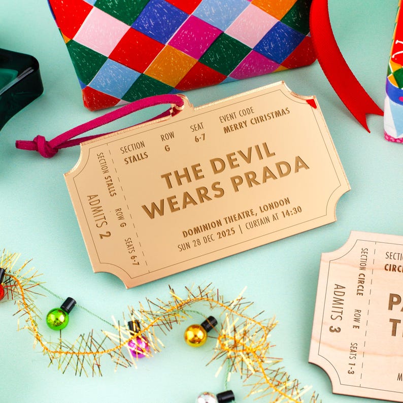 以下が含まれることがあります： 「THE DEVIL WEARS PRADA」とイベント詳細が記載された金色のチケット。ピンク色の紐で結ばれ、クリスマスの装飾に囲まれています。イベントはロンドンで開催されます。