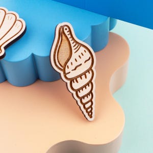 May include: Wooden seashell-shaped craft with a light brown interior and a darker brown outline. The seashell is positioned on a light peach-colored surface, with a blue background. The craft is likely for decorative purposes.