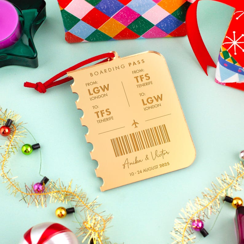 Op de afbeelding: Een goudkleurige boarding pass-vormige ornament met een rood touwtje. De pas bevat de tekst "BOARDING PASS", "FROM: LGW LONDON TO: TFS TENERIFE", en "Anika & Victor 10-24 AUGUST 2025".