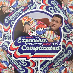 May include: A stack of colorful round stickers with a tie-dye pattern and a man's face. The stickers feature the text "The merch is so expensive because the music is so complicated".