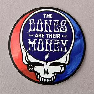 May include: A round sticker with a red, white, and blue swirl design. The center features a blue circle with white text that reads "The Bones Are Their Money". The text is in a stylized font. Below the text is a lightning bolt symbol. The blue circle is surrounded by a white outline and a white skull with a toothy grin.
