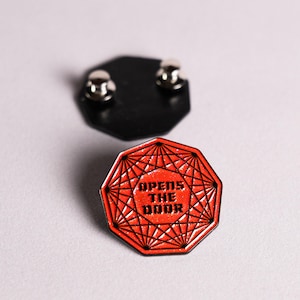 May include: An octagonal enamel pin featuring a vibrant red glitter background with black geometric line patterns and the text "OPENS THE DOOR" in bold black lettering. The back of the pin is black with two metal pin posts for secure attachment.