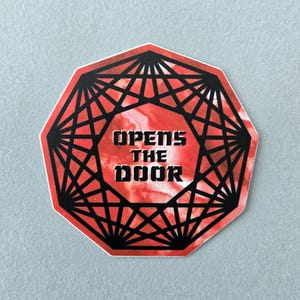 May include: A red and black sticker with the text "OPENS THE DOOR" in bold, white letters. The sticker has a geometric design with a red marbled background. The sticker is an octagon shape.