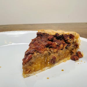 May include: A slice of pecan pie on a white plate. The pie has a golden brown crust and is filled with a rich, brown pecan filling. The filling is topped with a layer of caramel.