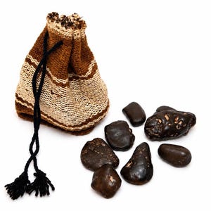 Puede incluir: Una pequeña bolsa de cordón tejida con rayas horizontales marrones y beige y cierre de cordón negro, acompañada de siete piedras oscuras de forma irregular, lisas y texturizadas, esparcidas sobre una superficie blanca.