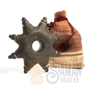 Puede incluir: Un objeto de piedra gris oscuro con forma de estrella, un orificio circular central y ocho protuberancias puntiagudas, acompañado de una pequeña bolsa de tela tejida en tonos marrón y beige atada con un cordón fino rojo oscuro y negro.