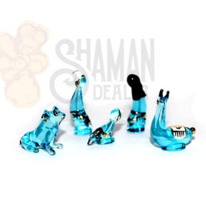 Puede incluir: Colección de pequeñas figuras de vidrio turquesa. El conjunto incluye un perro, una foca, dos figuras humanas estilizadas y una llama. Cada figura tiene detalles dorados y está sobre un fondo blanco.