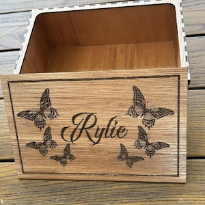 May include: A wooden box with a hinged lid. The lid is engraved with the name "Rylie" and several butterfly designs. The box is made of light brown wood.