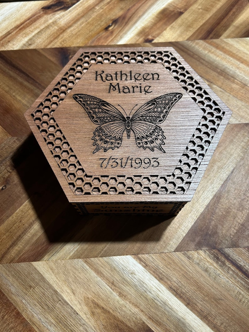 A hexagonal wooden box with a honeycomb pattern and a butterfly design. The box is engraved with the name 'Kathleen Marie' and the date '7/30/1993'.