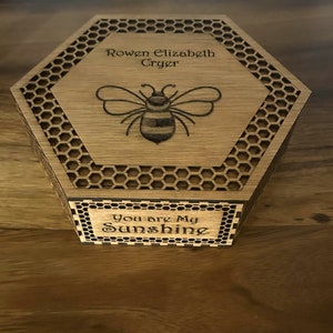 May include: Hexagon-shaped wooden box with a laser-etched bee design and the words "Rowen Elizabeth Cryer" on the lid. The box also features the phrase "You are My Sunshine" on the front. The box has a honeycomb pattern around the edges.