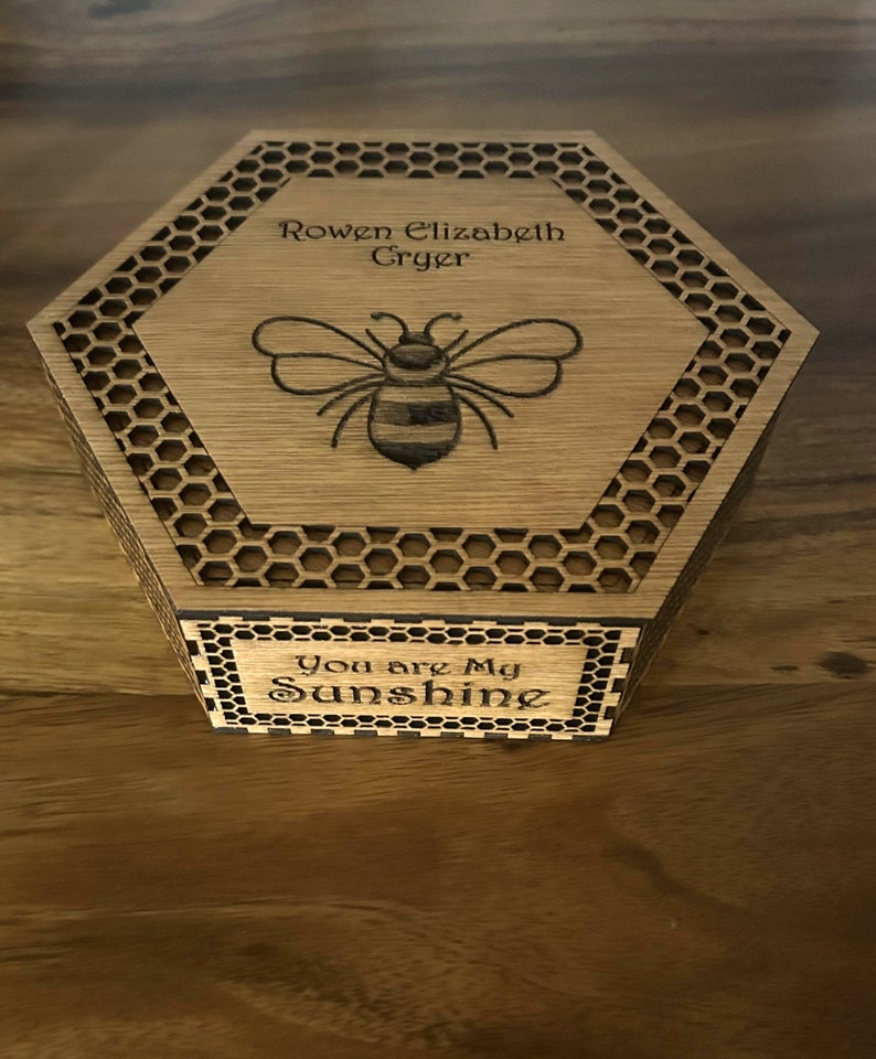 May include: Hexagon-shaped wooden box with a laser-etched bee design and the words "Rowen Elizabeth Cryer" on the lid. The box also features the phrase "You are My Sunshine" on the front. The box has a honeycomb pattern around the edges.