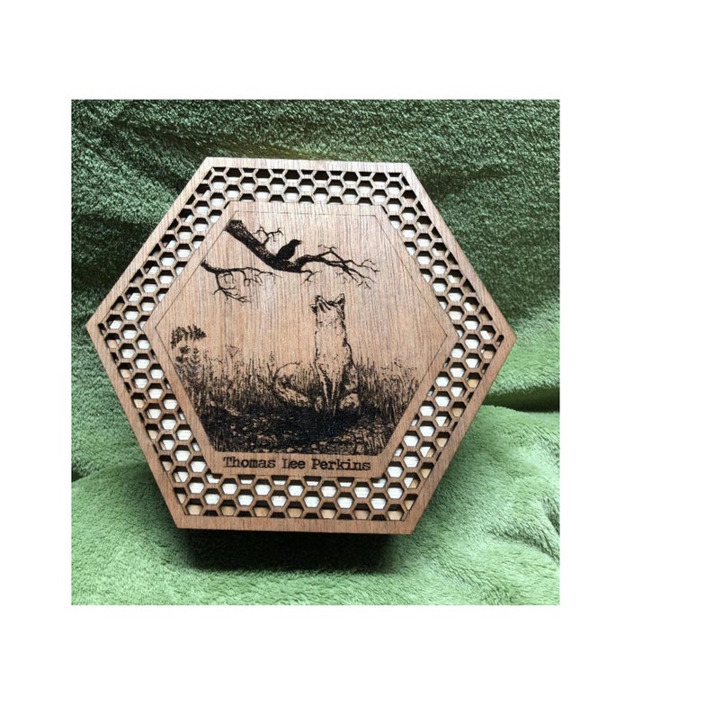 May include: A wooden, hexagonal box with a laser-etched design. The design features a fox looking up at a crow perched on a branch. The box has a honeycomb pattern around the central image and the text "Thomas Lee Perkins" at the bottom.