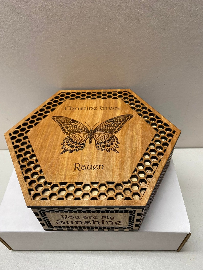 May include: A wooden, hexagonal box with a laser-etched butterfly design on the lid. The lid also features the names "Christine Grace" and "Rauen". The box has a honeycomb pattern around the edges and the phrase "You are My Sunshine" on the front.