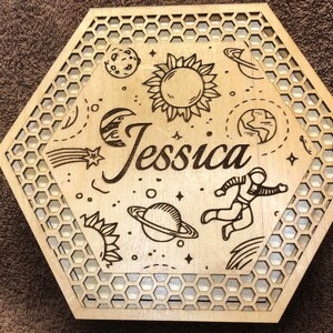 May include: A laser-cut wooden box with a hexagonal shape and a honeycomb pattern border. The box is decorated with space-themed illustrations, including planets, stars, a sun, and an astronaut. The name "Jessica" is prominently displayed.