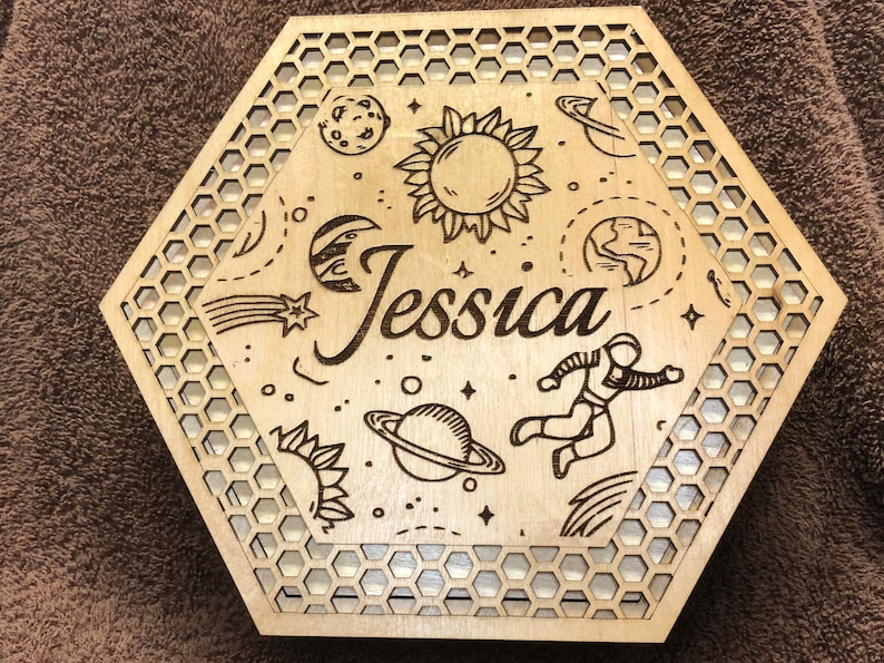 May include: A laser-cut wooden box with a hexagonal shape and a honeycomb pattern border. The box is decorated with space-themed illustrations, including planets, stars, a sun, and an astronaut. The name "Jessica" is prominently displayed.