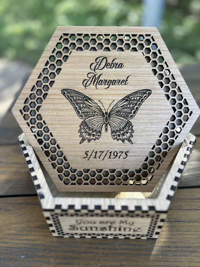 May include: A wooden, hexagonal box with a laser-cut butterfly design and the name "Debra Margaret" and the date "5/17/1975" engraved on the lid. The box has a honeycomb pattern around the edges and the words "You are My Sunshine" on the front.