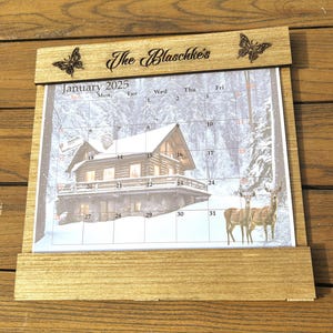 May include: Wooden desk calendar for January 2025, featuring a winter cabin scene and two deer. The calendar is framed in light wood and includes the text "The Blaschke's" and butterfly accents.