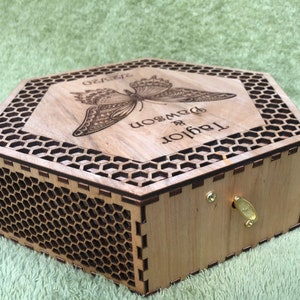 A hexagonal wooden box with a honeycomb pattern and a butterfly design engraved on the lid. The lid is open and the box has a brass keyhole and a brass key. The lid is engraved with the text 'Taylor & Dawson' and the text 'Forever'.