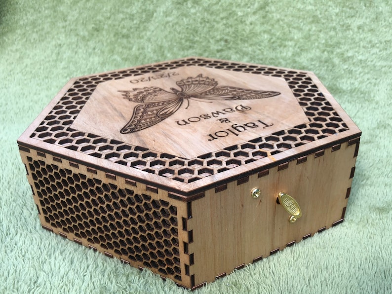 A hexagonal wooden box with a honeycomb pattern and a butterfly design engraved on the lid. The lid is open and the box has a brass keyhole and a brass key. The lid is engraved with the text 'Taylor & Dawson' and the text 'Forever'.