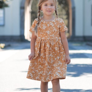 以下が含まれることがあります： 白の花柄のマスタード色のドレス。ドレスは半袖で、ギャザーのスカートが付いています。女の子は建物の前でドレスを着ています。