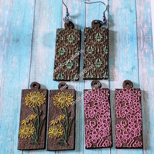 Puede incluir: Cuatro pares de pendientes de madera con diferentes diseños. Dos pares presentan un patrón floral verde y marrón, un par tiene un diseño de flores amarillas y el último par tiene un diseño floral rosa.