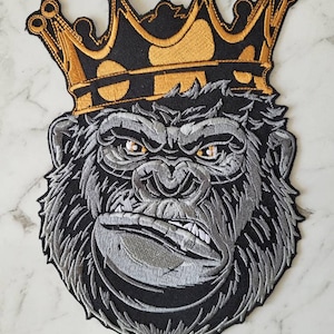 May include: A black and silver embroidered patch of a gorilla wearing a gold crown. The gorilla has a fierce expression and is looking directly at the viewer.