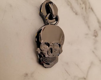Puede incluir: Un tirador de cremallera de metal negro con un diseño de calavera. La calavera está detallada con cuencas de los ojos, una nariz y una boca.