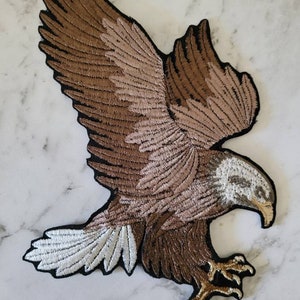 Może przedstawiać: Naszywka z haftowanym brązowym i białym orłem z czarną obwódką. Orzeł jest w locie z rozpostartymi skrzydłami. Głowa orła jest zwrócona w prawo, a jego dziób jest otwarty.