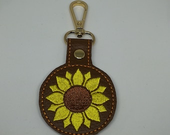 Puede incluir: Un llavero de cuero marrón con un cierre de metal dorado y un girasol amarillo bordado.