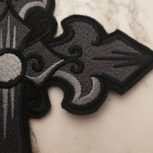 May include: A black and gray embroidered cross patch with a white center circle. The cross has a detailed design with intricate swirls and lines.