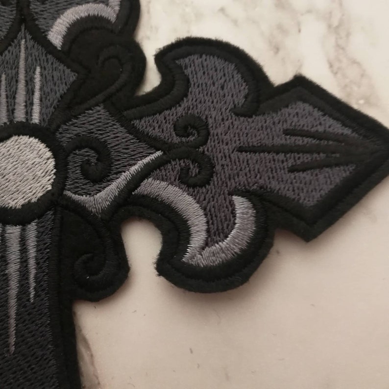 May include: A black and gray embroidered cross patch with a white center circle. The cross has a detailed design with intricate swirls and lines.